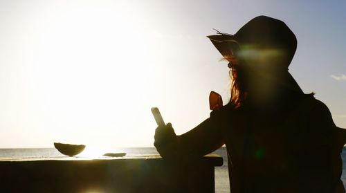 Rear view of silhouette woman sitting at table with sea in background against sky