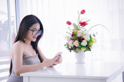 Woman using mobile phone by flower vase at home