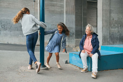 Smiling grandmother looking at playful granddaughters spinning around pole at playground