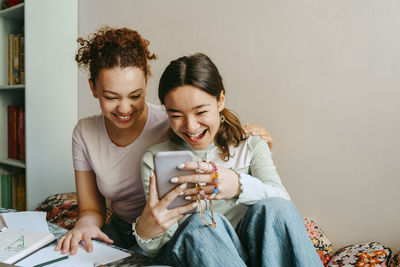 Cheerful young woman sharing smart phone with teenage friend at home