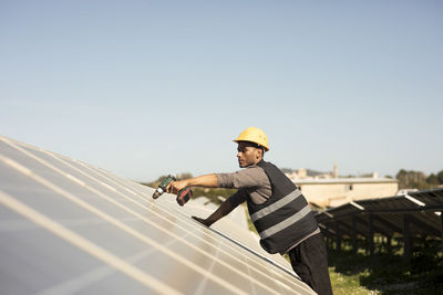 Side view of male maintenance engineer drilling on solar panels while working at power station