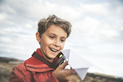 Smiling boy holding paper airplane while standing against cloudy sky