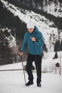 Teenager in a white cap with a pompom and a blue jacket climbs a mountain using trekking poles 