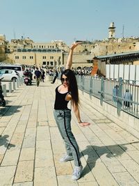 Full length portrait of young woman standing in city against sky