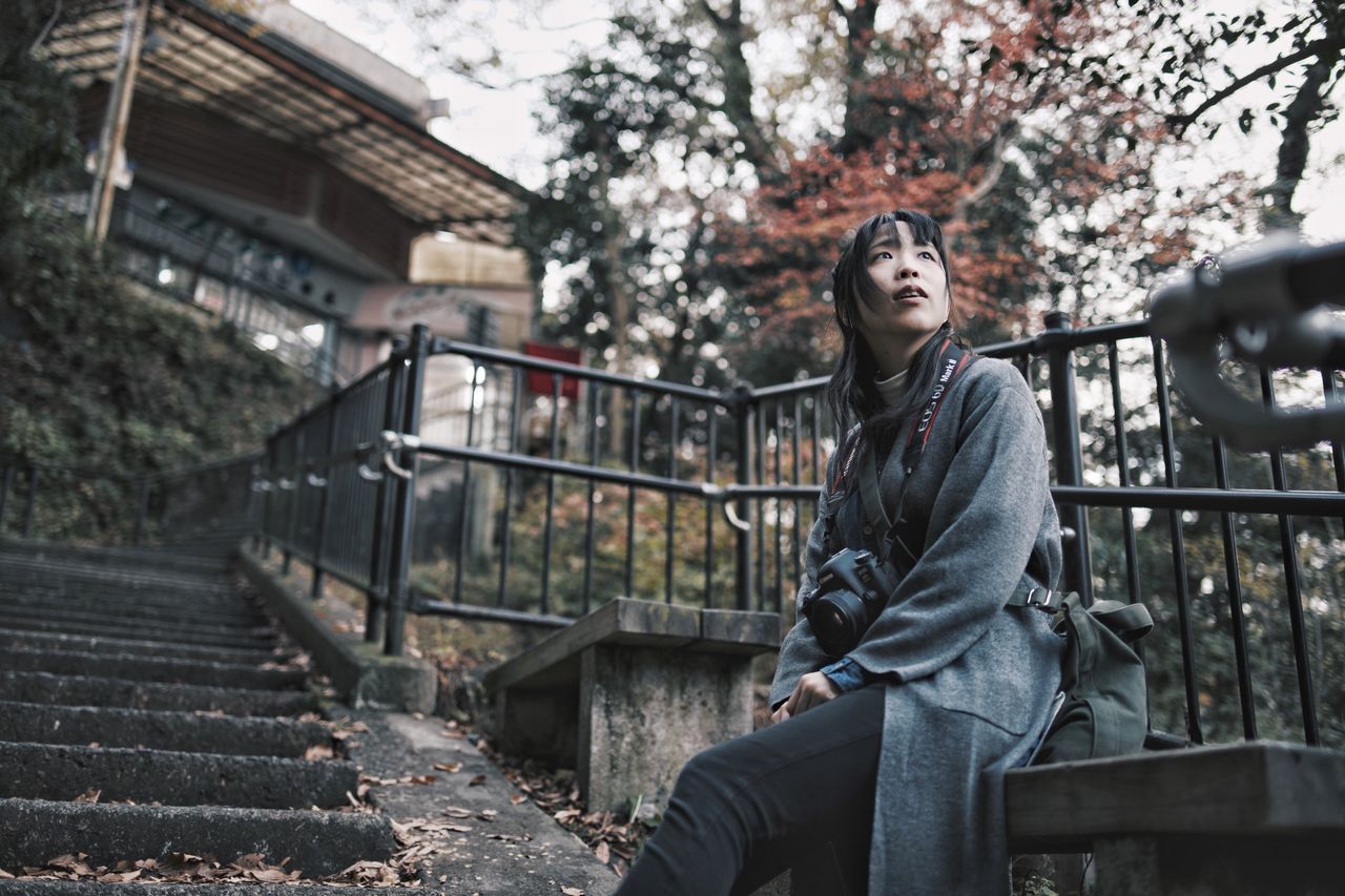 one person, real people, architecture, leisure activity, lifestyles, young adult, three quarter length, built structure, tree, railing, sitting, focus on foreground, casual clothing, young women, nature, day, front view, clothing, outdoors, contemplation, hairstyle, warm clothing