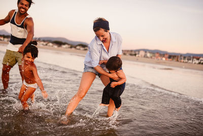 Happy multiracial family with children playing together in shallow water of sea near coast against residential buildings on summer day