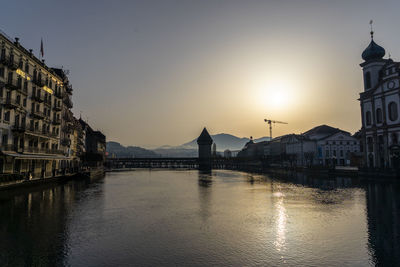 Bridge over river by buildings against sky during sunset