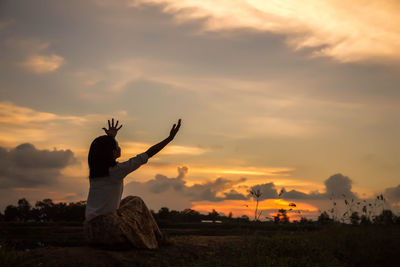 Woman with arms raised sitting on field against sky during sunset