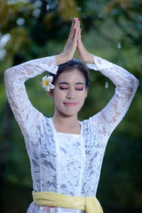 Portrait of young woman with arms raised standing against trees