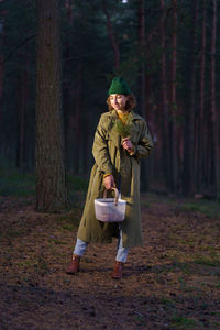 Young woman in hipster hat and trendy coat smiles standing with basket at dark trees