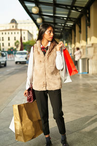 Calm hispanic female with shopping bags strolling on walkway along road with car on street of city looking away