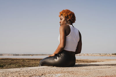Back view of african american female in white top and leather pants sitting on shore of sea and looking away in sunny daylight