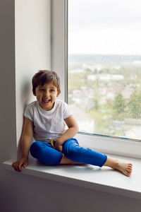 Boy in a white tank top and blue pants sits on a window in rainy weather