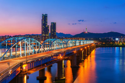 Illuminated bridge over river by buildings against sky at night