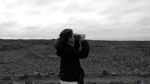 Woman looking through binoculars on field against sky