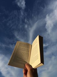 Low angle view of hand holding book against sky