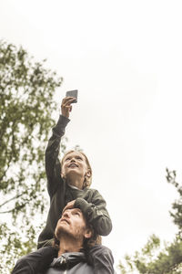 Low angle view of girl photographing through mobile phone while sitting on father's shoulders against sky