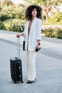 Full length hispanic businesswoman in white suit standing on pavement near suitcase and looking away over shoulder during business trip on summer day