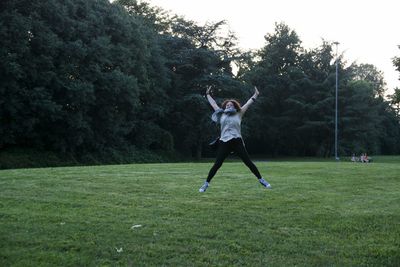 Playful woman jumping on grassy field at public park against trees