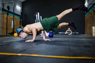 Shirtless young man balancing on floor while woman resting in background at gym