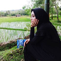 Thoughtful woman in hijab sitting against farm