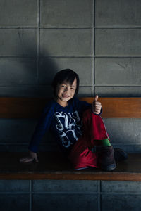 Full length portrait of smiling boy showing thumbs up sign while sitting on bench