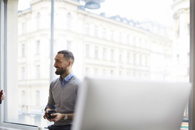 Smiling businessman holding cup while sitting against window at office