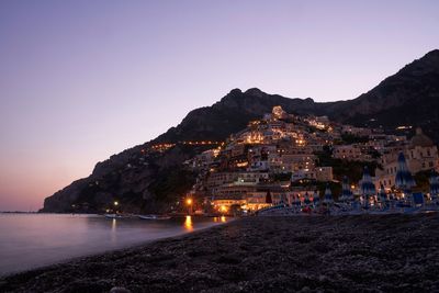 Illuminated buildings by sea against clear sky at dusk