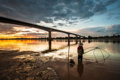 Man fishing in river against bridge during sunset