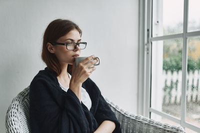 Beautiful young woman looking through window at home