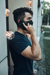 Young man standing in front of restaurant talking on phone using smartphone wearing the face mask