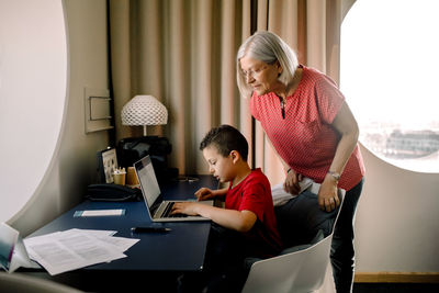 Grandmother looking at grandson using laptop while sitting by table in hotel room