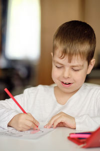 Cute and smiling cucasian boy draws with colored pencils while sitting at the table. 