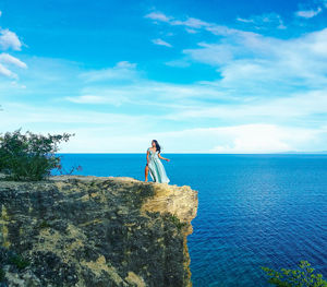 Woman standing on cliff by sea against blue sky