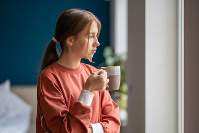 Upset unhappy teenage girl holding tea cup looking out window