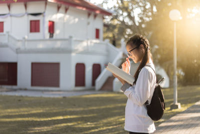 Side view of young woman holding book while standing on footpath