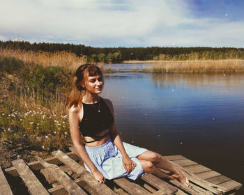 Portrait of smiling young woman sitting by lake against sky