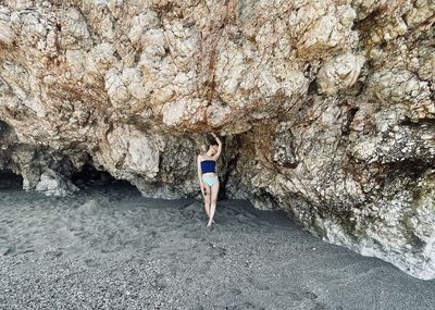 Woman in blue swimsuit stand under huge rock at silent beach