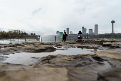 People standing on shore against cloudy sky