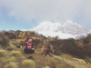 Portrait of man sitting by yak on mountain against sky