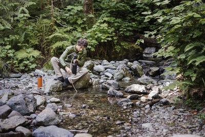 Hiker filling water bottle while sitting on rock at lakeshore in forest