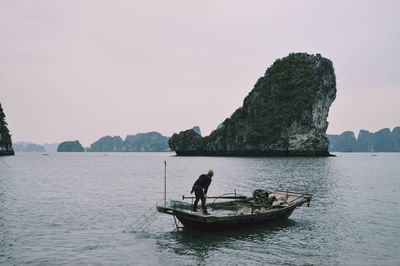 Man standing on boat in halong bay with rocky mountains in background against clear sky