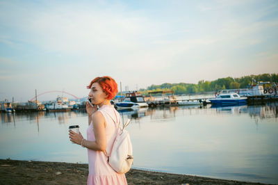 Mid adult woman talking on mobile phone while standing by river against sky during sunset