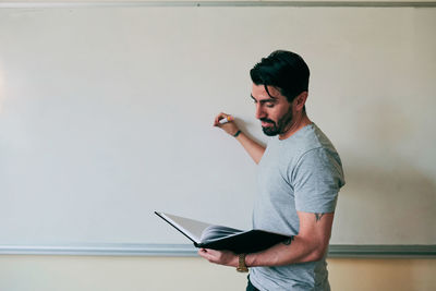 Confident male teacher holding book while writing on whiteboard