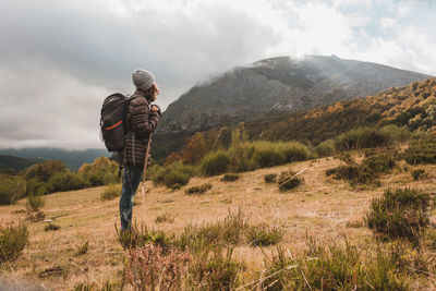 Side view of hiker with backpack standing on mountain against cloudy sky