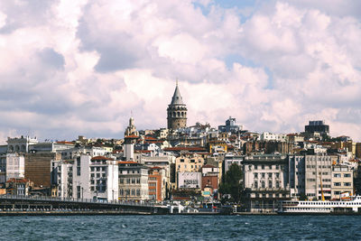 Galata tower amidst buildings in front of river against cloudy sky in city