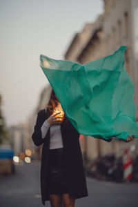 Woman with green scarf and illuminated jar standing on city street at dusk