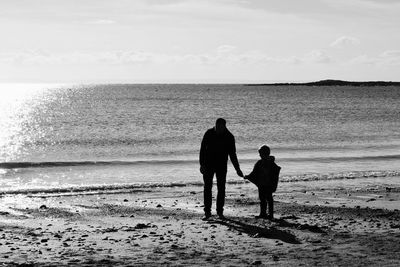 Silhouette father and son standing on shore at beach against sky
