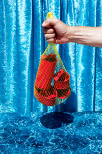 Crop unrecognizable person with mesh bag with assorted aluminum red tins from preserves on blue background in studio