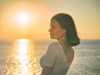 Side view of young woman standing at beach against sky during sunset